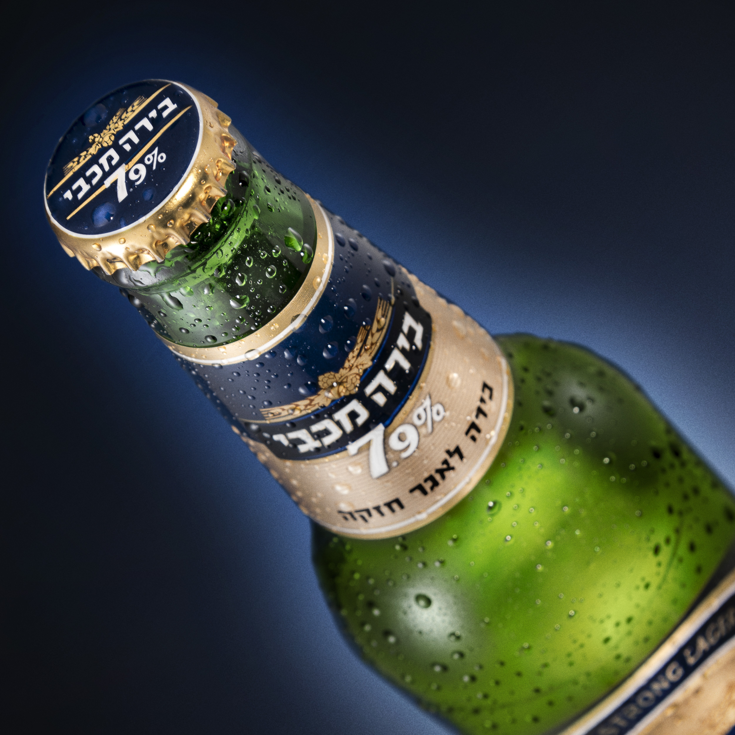 בקבוק זכוכית בירה מכבי 7.9% בקבוק ירוק עם תוית כחול זהוב על רקע מעברי כחול שחור צולם עבור משרד פרסום CLIMEX ע'י סטודיו רייס