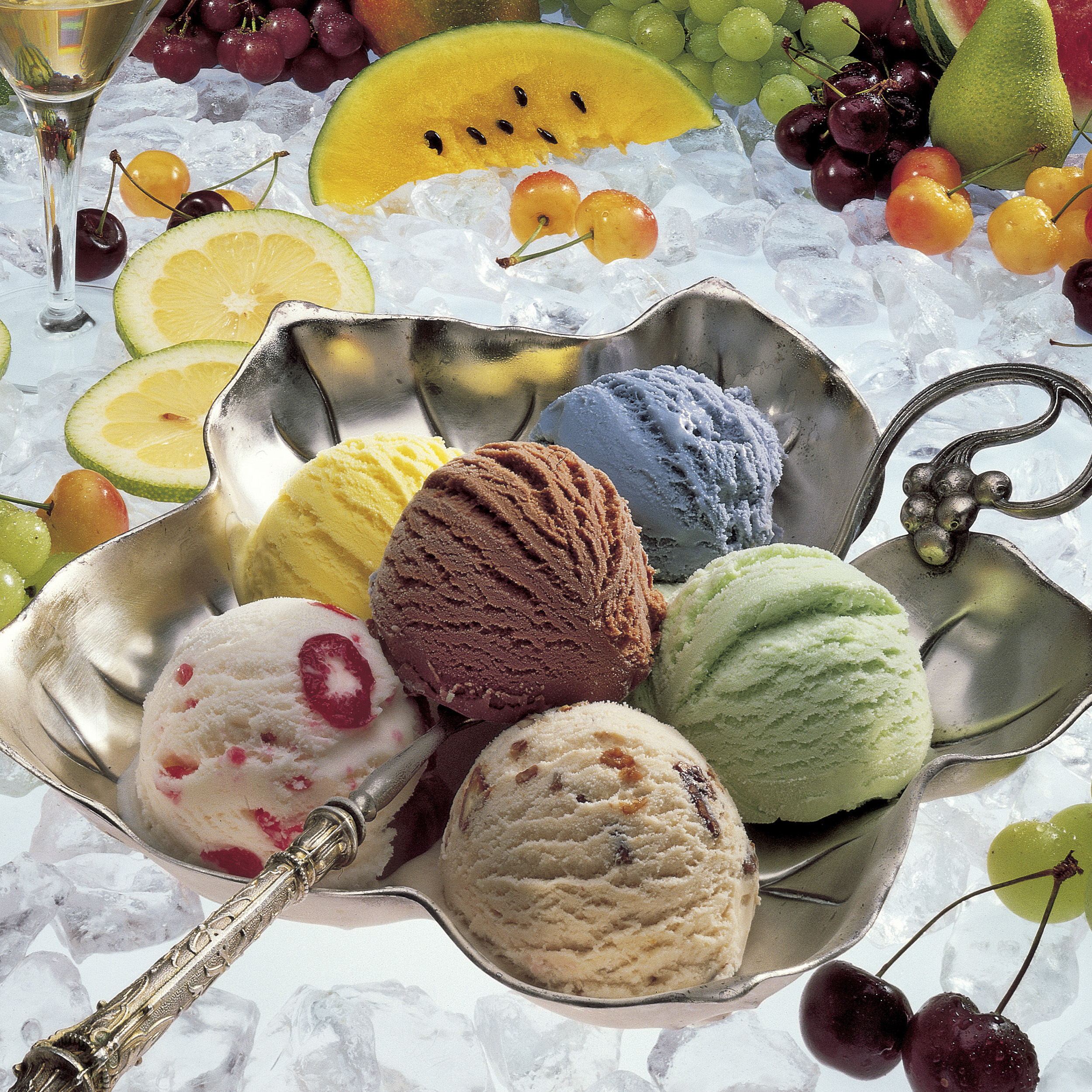 כלי כסף עם 6 כדורי גלידה בצבעים שונים בכלי כסף על משטח קרח לבן עם קומפוזיציה של פירות קיץ רעננים. סטיילינג של נורית ברניצקי צילום של סטודיו רייס