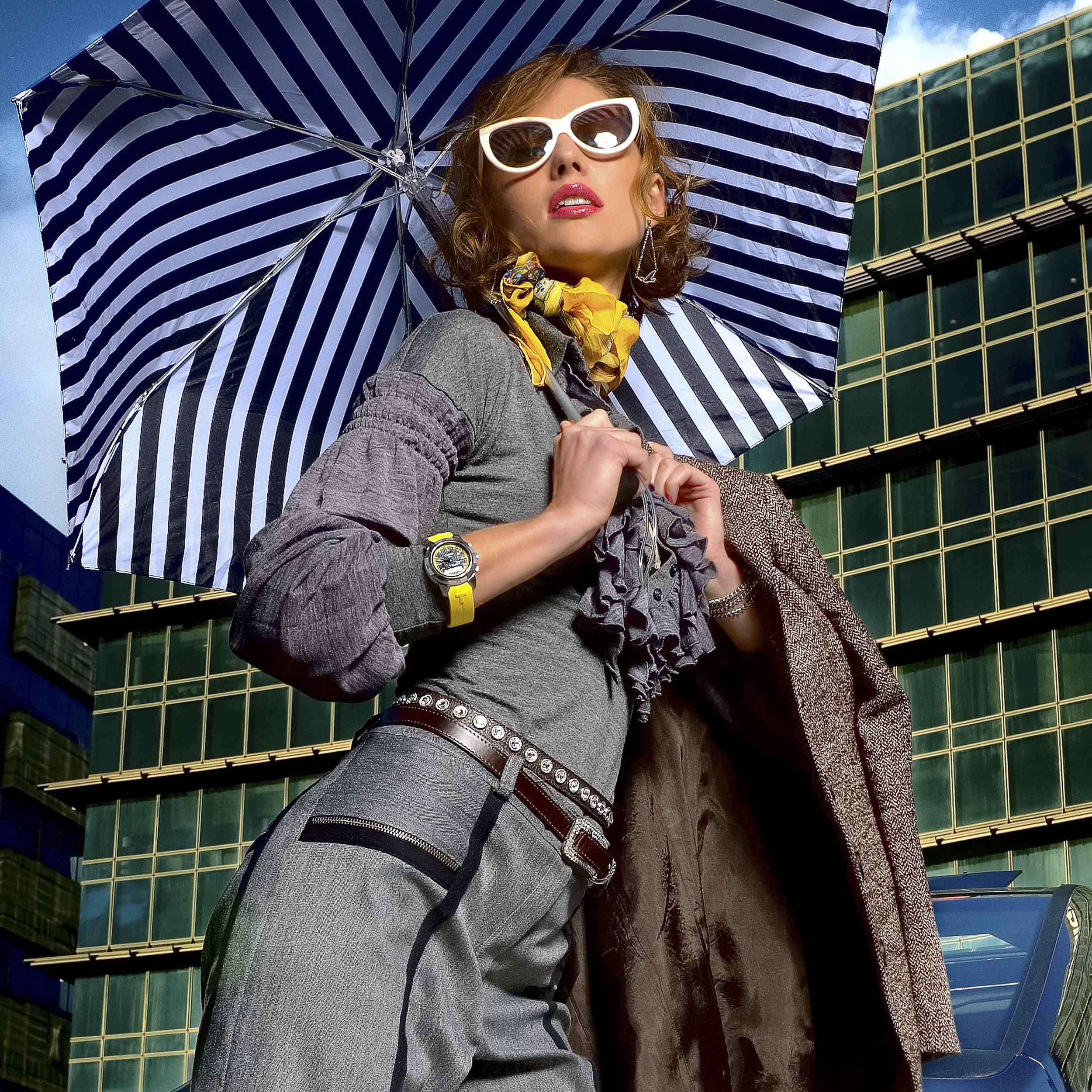 דוגמנית בחליפה נשית אפורה ג'קט וחגורה חומים עם משקפי שמש לבנים שעון עם רצועה צהובה של צהוב עגילים וצמיד כסופים ליפסטיק ורוד עם שימשיה שחור לבן על רקע אורבני בניין משרדים ליד רכב מרצדס עם מזוודה בורדו צולם ע'י סטודיו רייס