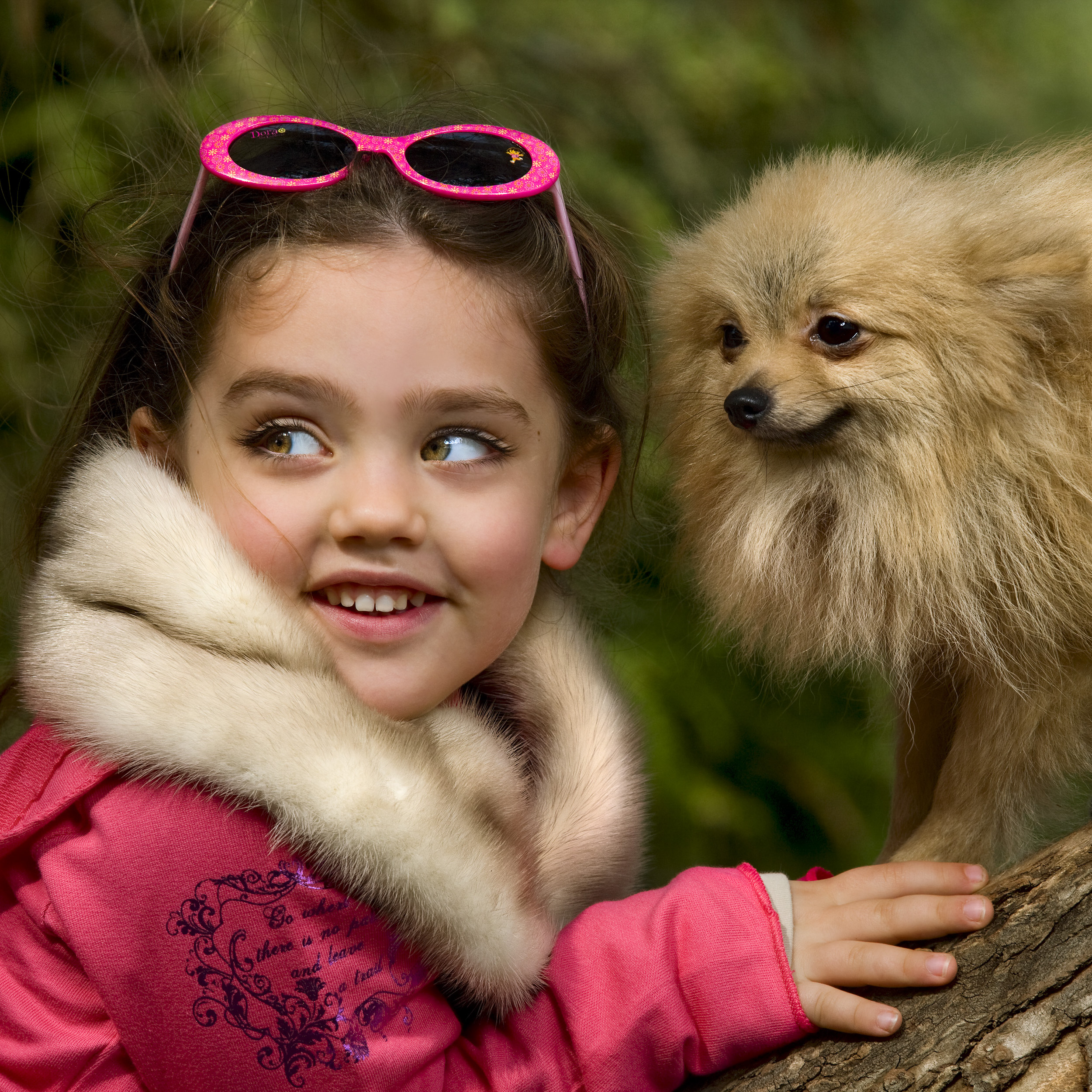 ילדה במעיל ורוד ומשקפי שמש נשענת על גזע עץ עם כלבלב קטן על רקע ירוק מטושטש.