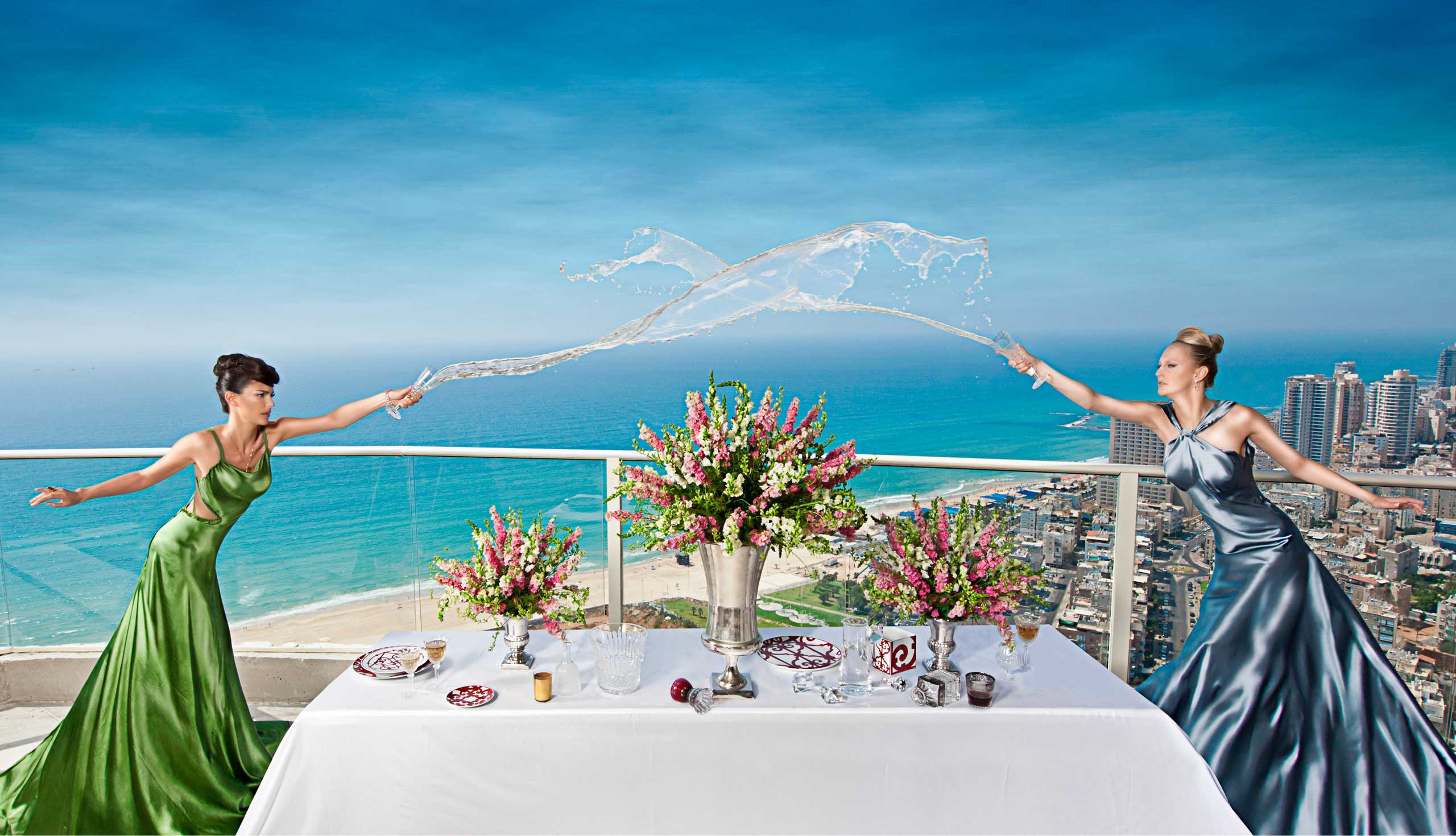 שתי דוגמניות בשמלת ערב סאטן כחולה וירוקה שופכות שמפניה אחת לכיוון השניה מעל שולחן עם סידורי פרחים ברקע נוף מהקומה ה29 במגדל על חוף בת ים